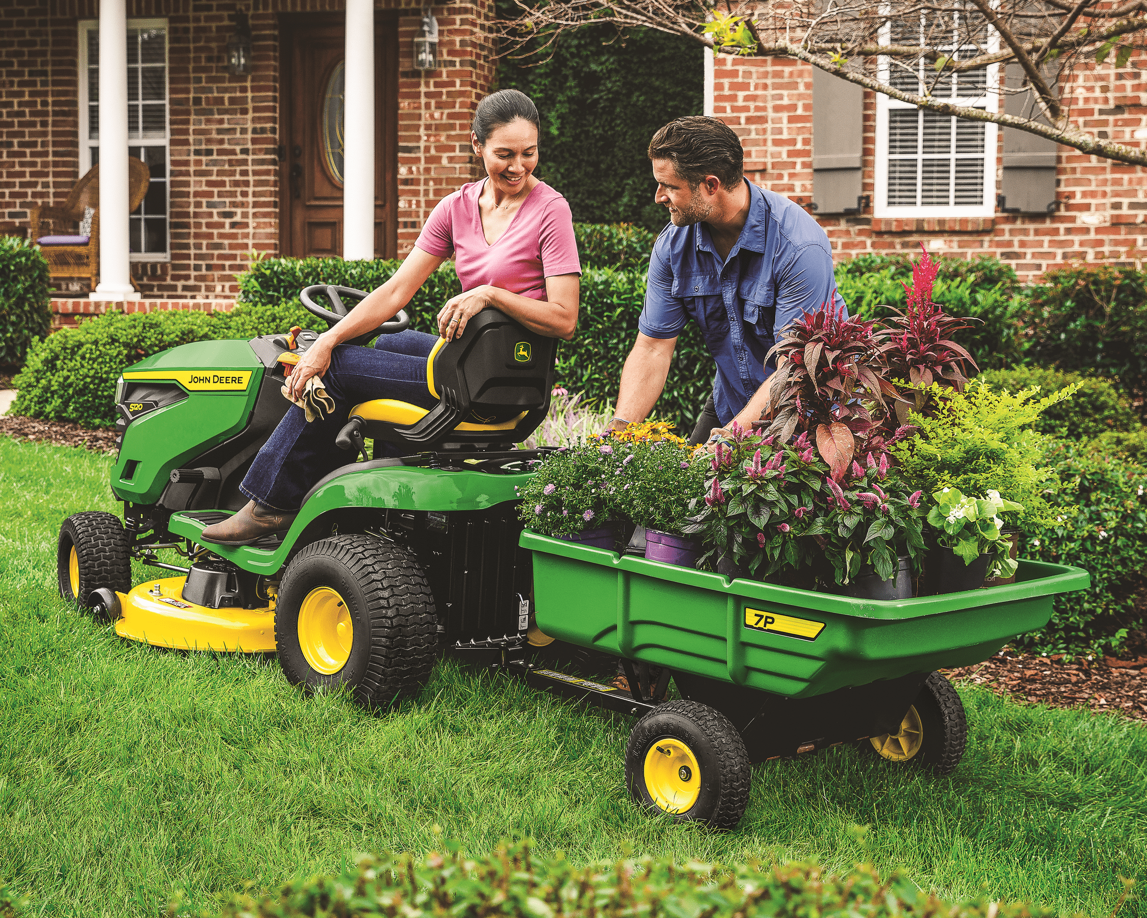 A couple is doing gardening work using a John Deere s120 lawn tractor with 7p cart.